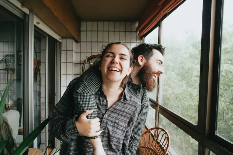 Two people laughing together on a balcony, one leaning on the other’s shoulder
