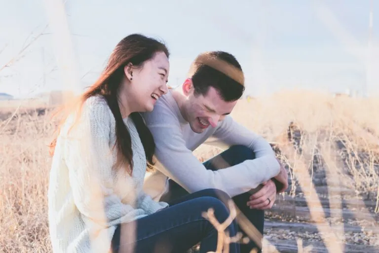 A couple sitting together in tall grass, laughing, photographed through the reeds