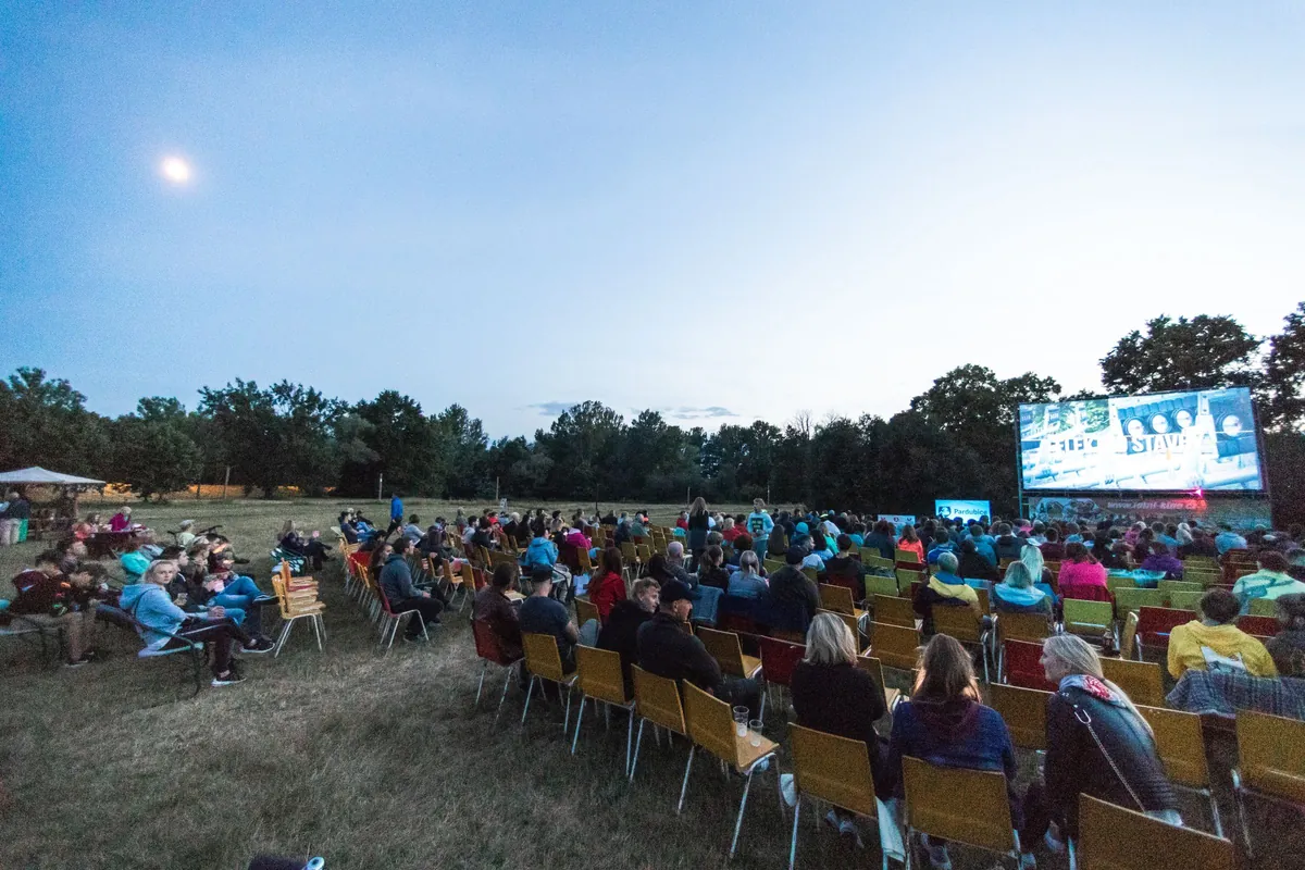 Rows of people in camp chairs watching a film on a large outdoor screen at dusk