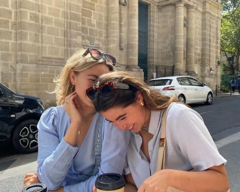Two friends leaning into each other laughing on a city street, holding takeaway coffees