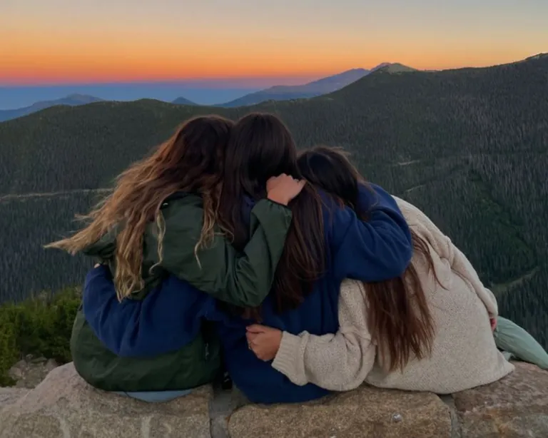 Four friends sitting with their arms around each other on a mountainside at sunset