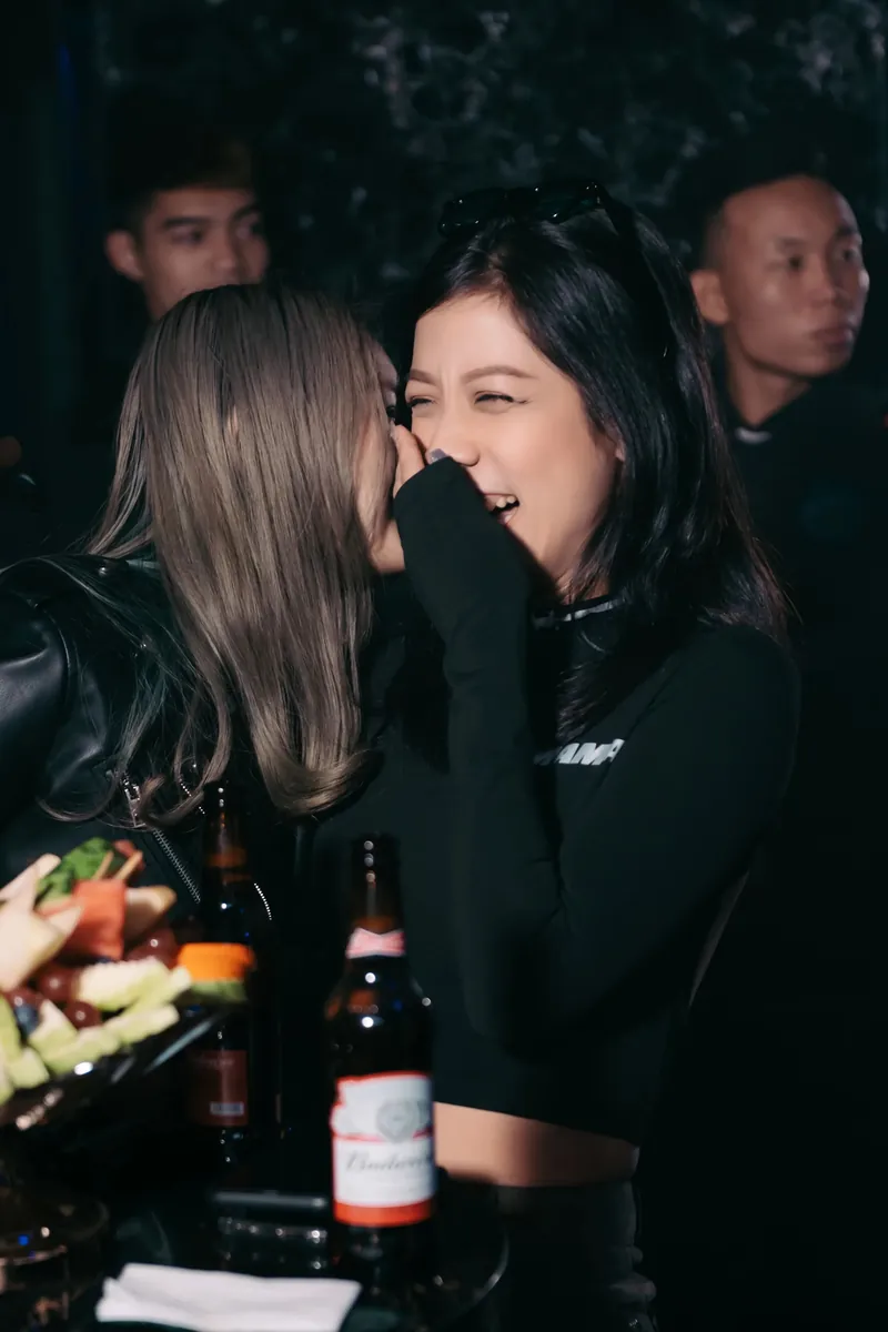 Two friends leaning in close at an evening event, one laughing at something the other just said