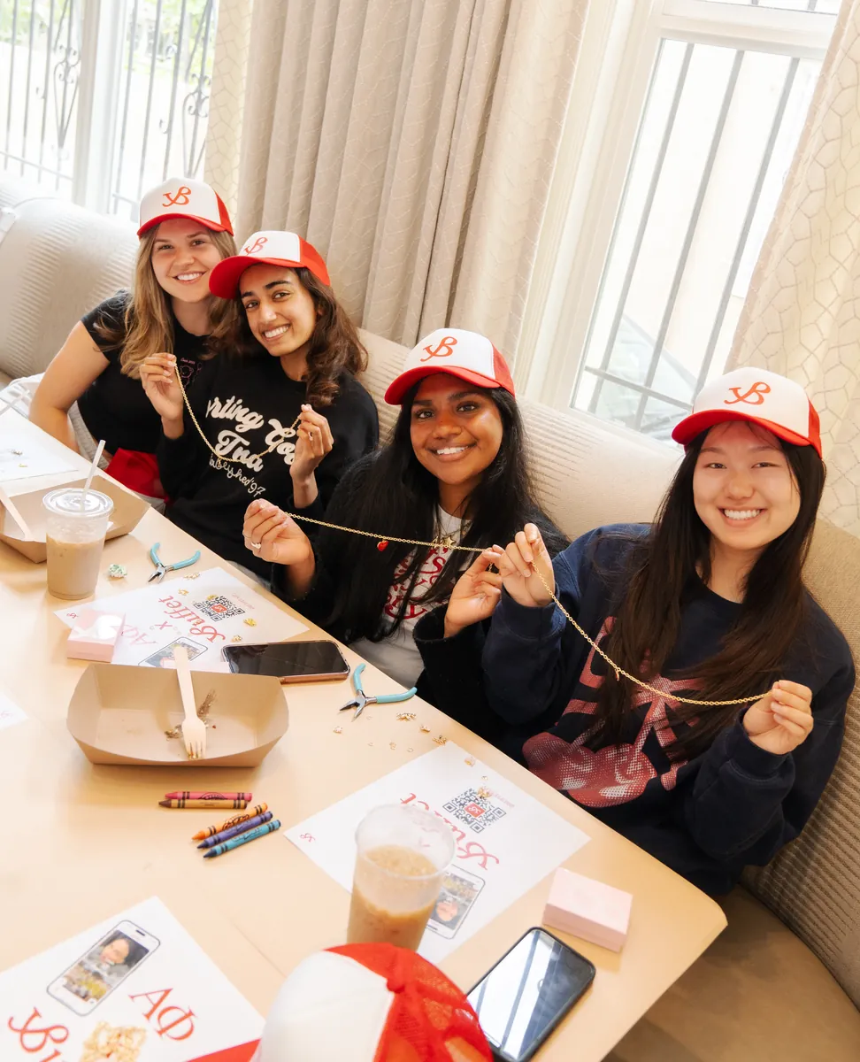 Five young women in red Buffet caps around a table, making friendship bracelets with iced coffees and craft supplies in front of them