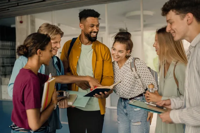 A group of friends standing together in a hallway, talking and looking at a tablet