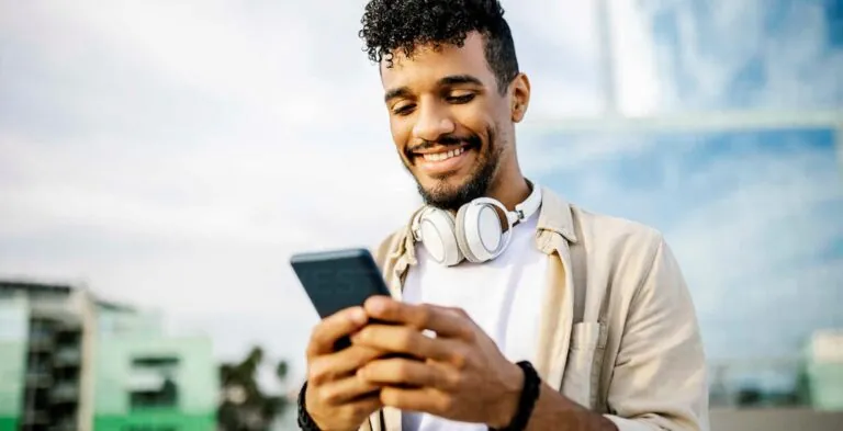 A man with headphones around his neck smiling at his phone outdoors
