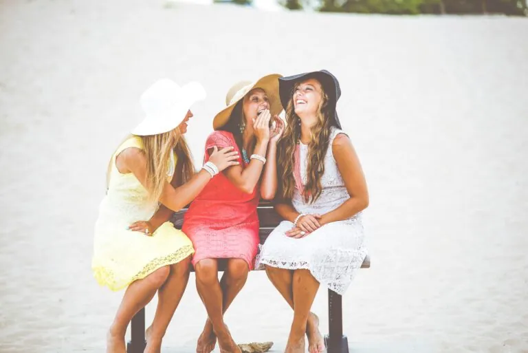 Four women in sun hats sitting on a bench at the beach, laughing together