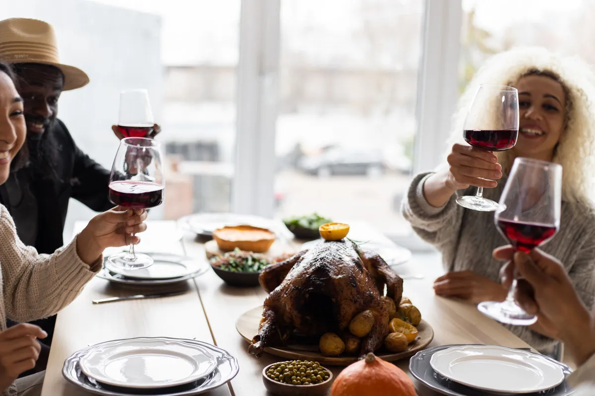 Four friends raising glasses of red wine over a roast turkey and a table of side dishes by a bright window