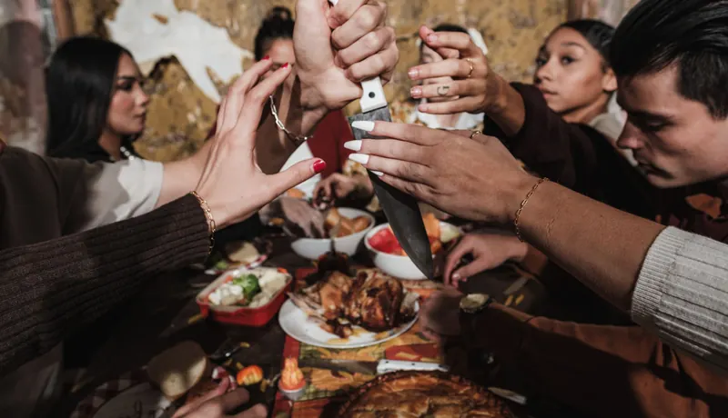 Six friends' hands stacked on the handle of a carving knife above a Thanksgiving turkey and a pie