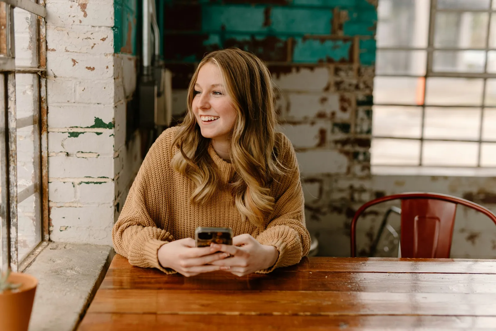 A woman smiling at her phone while sitting at a wooden table beside a window