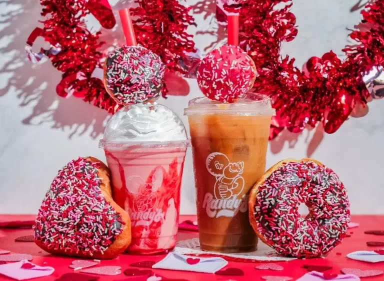 Two heart-shaped donuts covered in red and white sprinkles beside a whipped-cream shake and an iced coffee on a red table