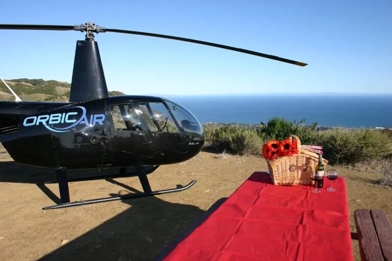 An Orbic Air helicopter on a clifftop above the ocean, with a red carpet and picnic basket laid out beside it