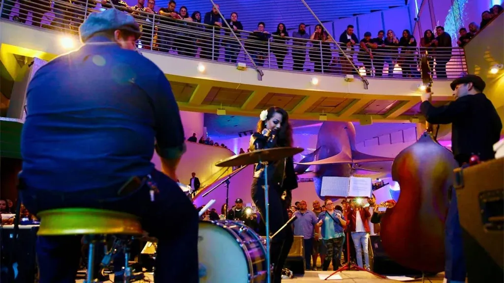 A band playing upright bass and drums under blue and purple light while a crowd watches from the balcony above