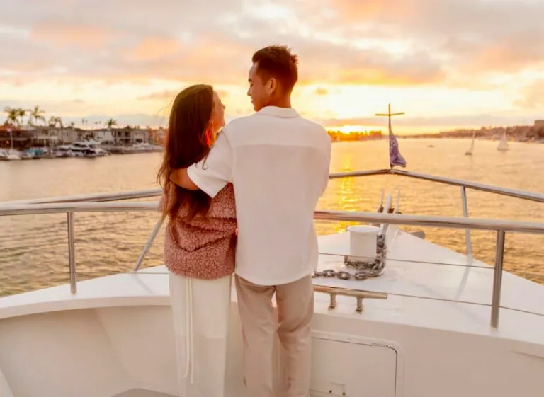 A couple standing at the railing of a yacht, watching the sun set over the marina