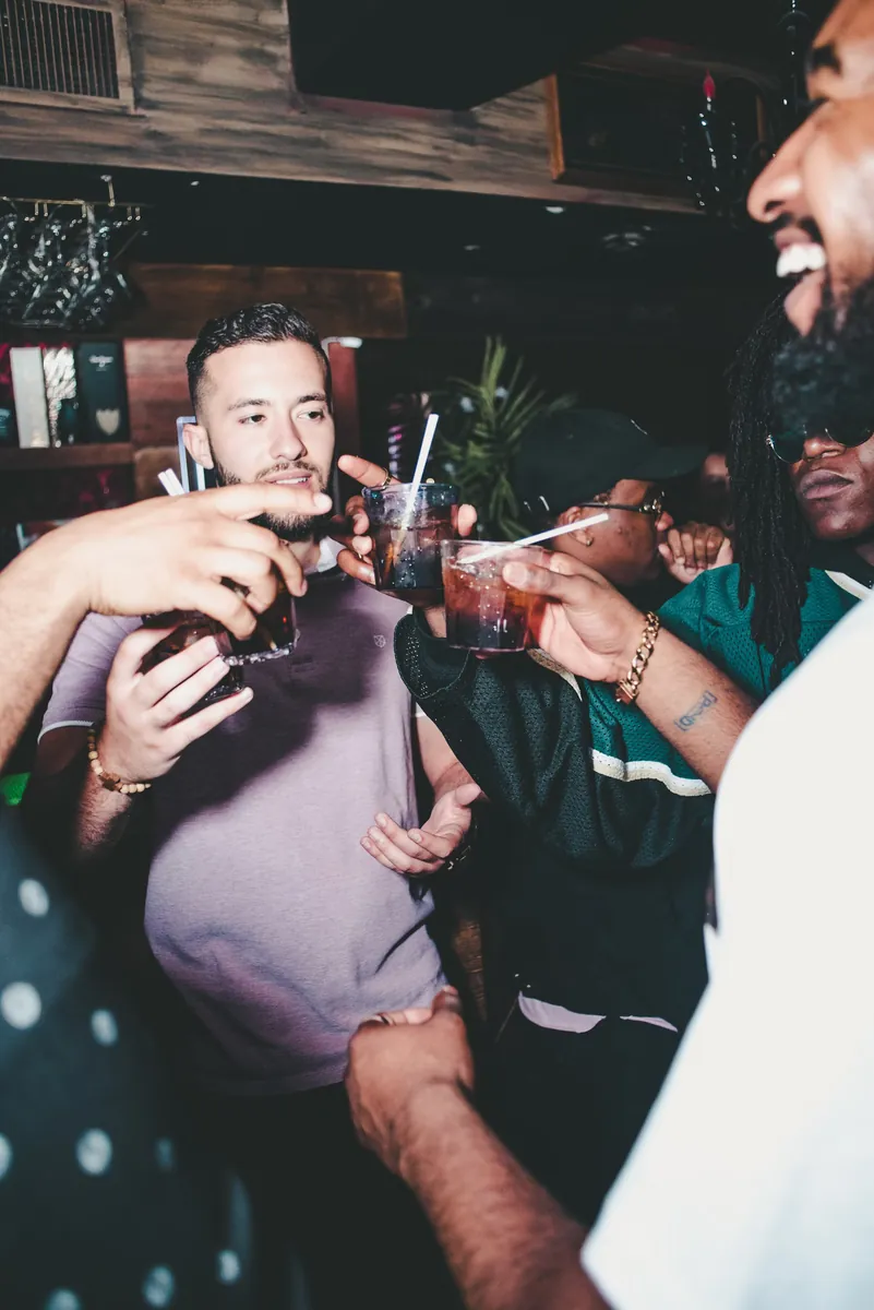 A group of friends leaning in together, laughing and touching cups at a crowded bar