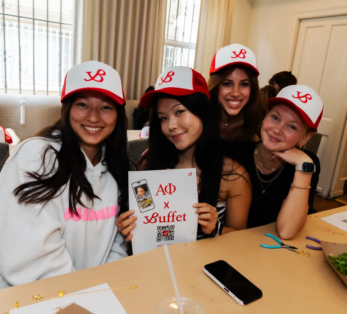 Four young women in red Buffet caps around a table, one holding up an Alpha Phi x Buffet flyer