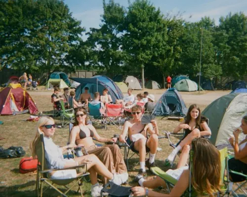 Festival-goers relaxing in camping chairs in front of tents on a sunny campsite